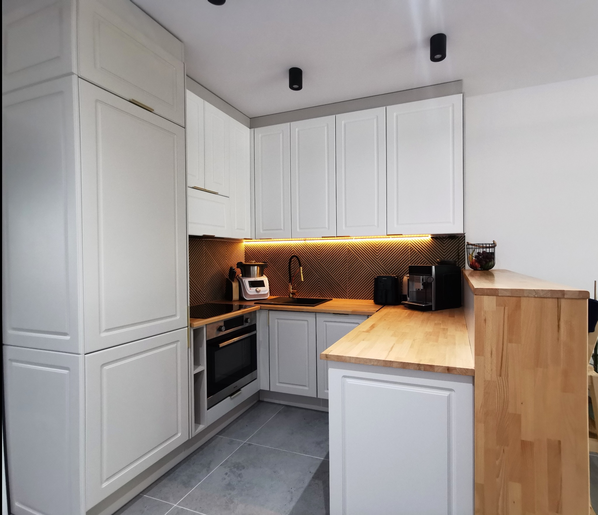Jasna kuchnia z białymi szafkami i drewnianym blatem. Uwagę przykuwa złota armatura i oświetlenie LED pod szafkami, które podkreśla dekoracyjny panel ścienny.