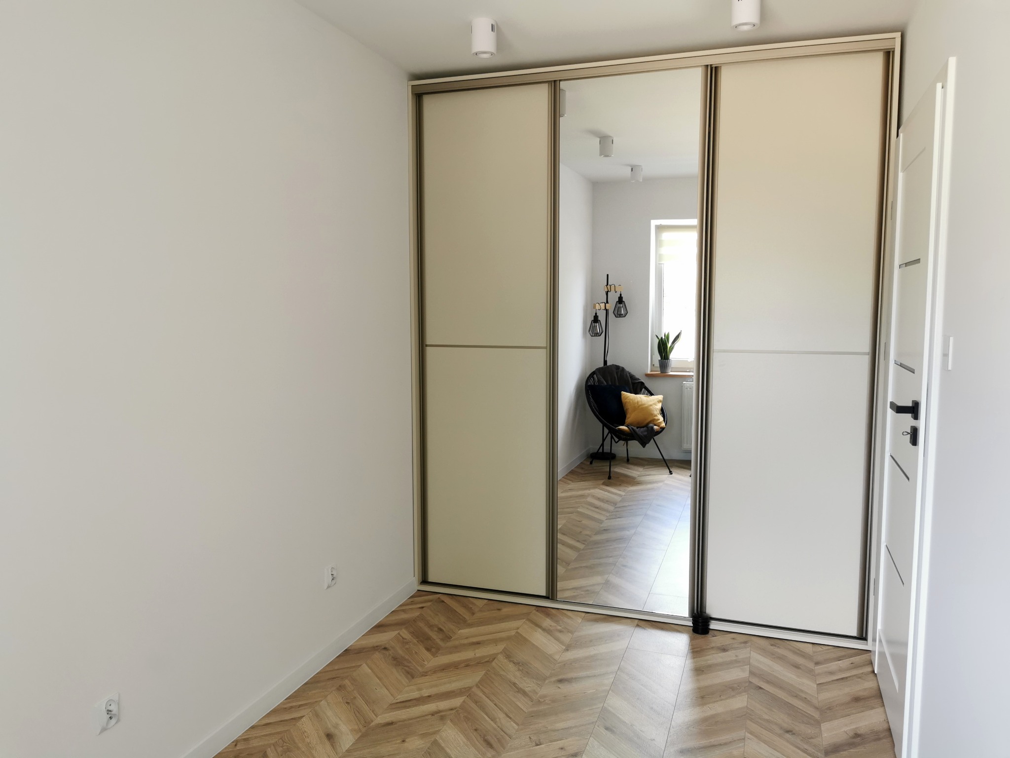 Jasna szafa przesuwna z lustrem w minimalistycznym wnętrzu. Parkiet w jodełkę, białe ściany i drzwi. W lustrze odbija się krzesło i roślina w doniczce.