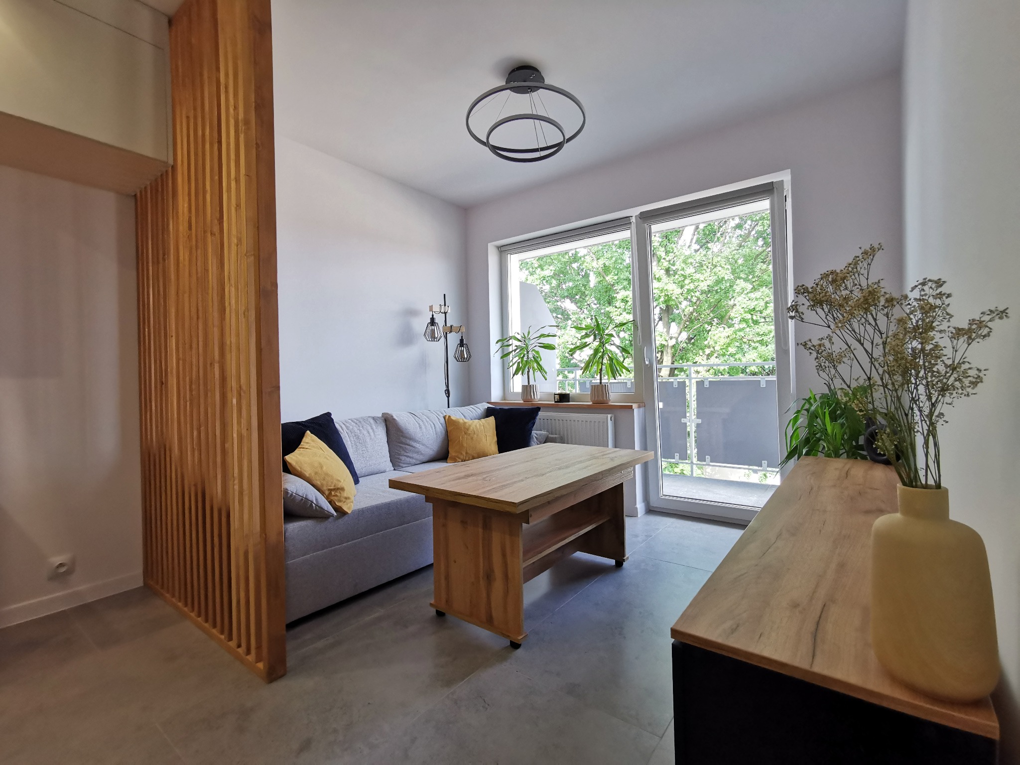 Jasne wnętrze z drewnianym ażurowym przepierzeniem, sofą z poduszkami, stolikiem kawowym i komodą. Za oknem zieleń. Nowoczesny design.