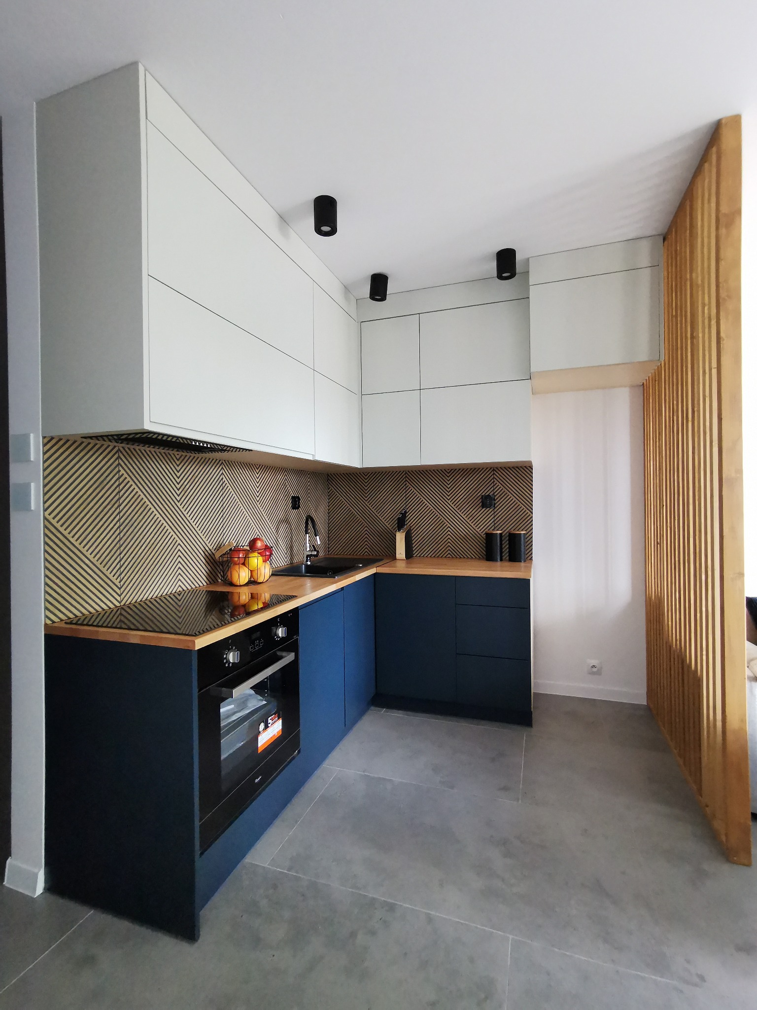 Nowoczesna kuchnia z białymi szafkami górnymi i granatowymi dolnymi, drewnianym blatem i geometrycznym wzorem na ścianie. Widoczny piekarnik i owoce w koszyku.