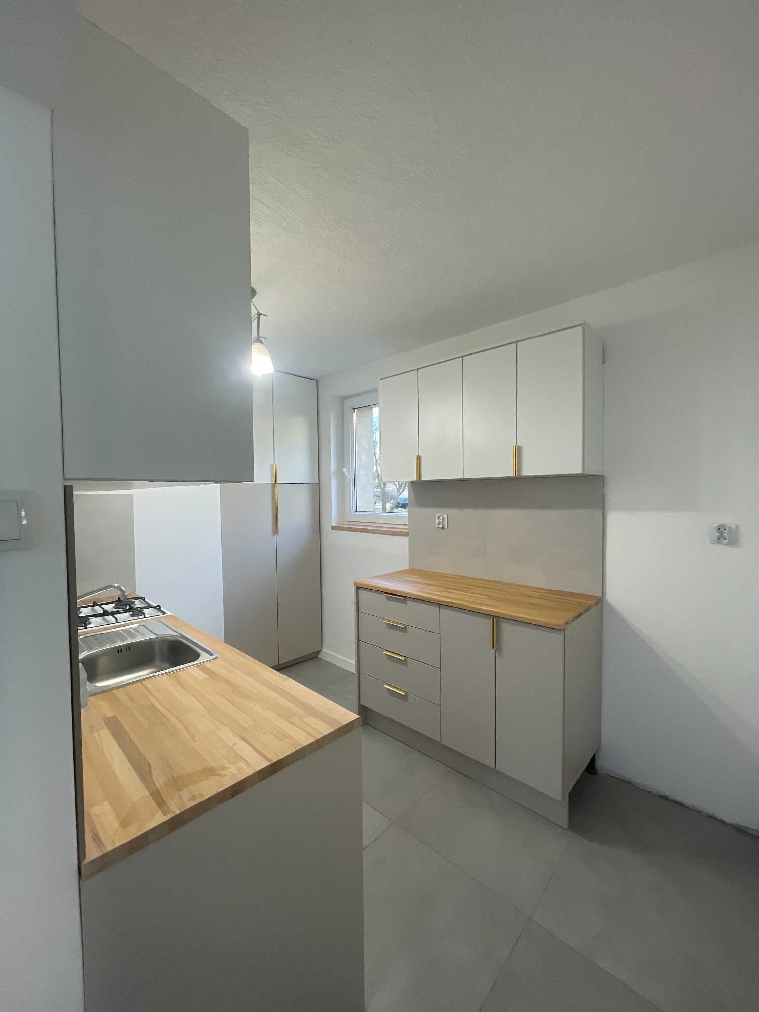 Jasna kuchnia z drewnianym blatem i złotymi uchwytami. Minimalistyczny design, szare płytki podłogowe. Widok na okno i szafę. Nowoczesne wykończenie.