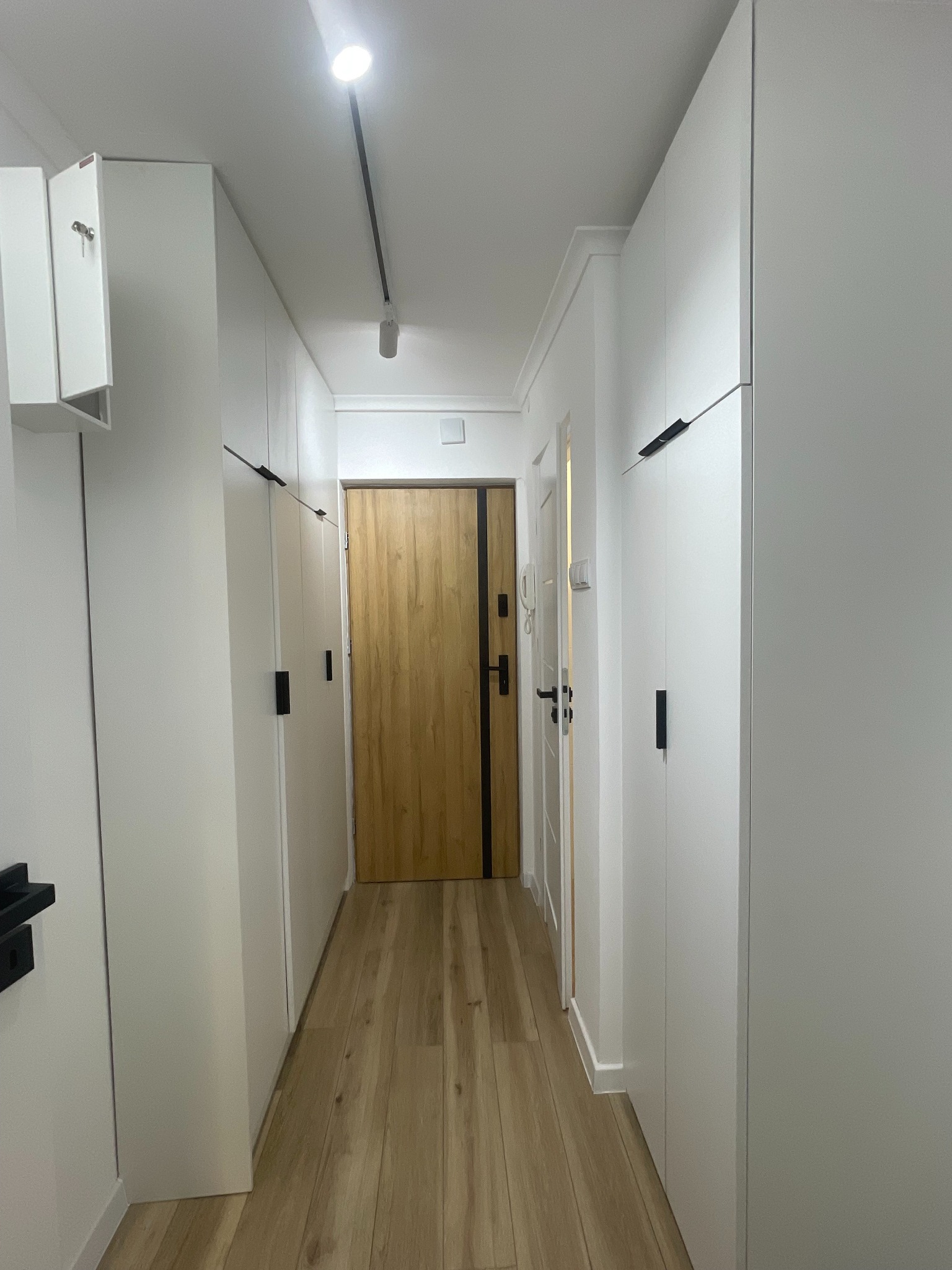 Białe szafy wnękowe z czarnymi uchwytami, zabudowa od podłogi do sufitu w wąskim korytarzu z drewnianą podłogą i drzwiami. Minimalistyczny design.