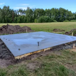 Wylana, świeża płyta fundamentowa pod budynek na tle zielonej łąki i lasu, widoczne drewniane szalunki i wystające rury kanalizacyjne