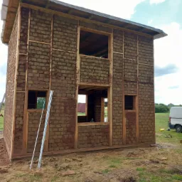 Niedokończony dom z cegły mułowej z drewnianą ramą, widoczne okna i dach, teren budowy z trawą i pojazdem w tle.