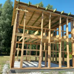 Konstrukcja z drewna w trakcie budowy, widoczne słupy, belki stropowe i zadaszenie, na betonowej wylewce, w tle drzewa i błękitne niebo.