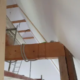 Poddasze w trakcie prac wykończeniowych: widoczne ocieplenie skośnego dachu, drewniana konstrukcja nośna z dekoracyjnymi rzeźbieniami oraz rusztowanie malarskie.