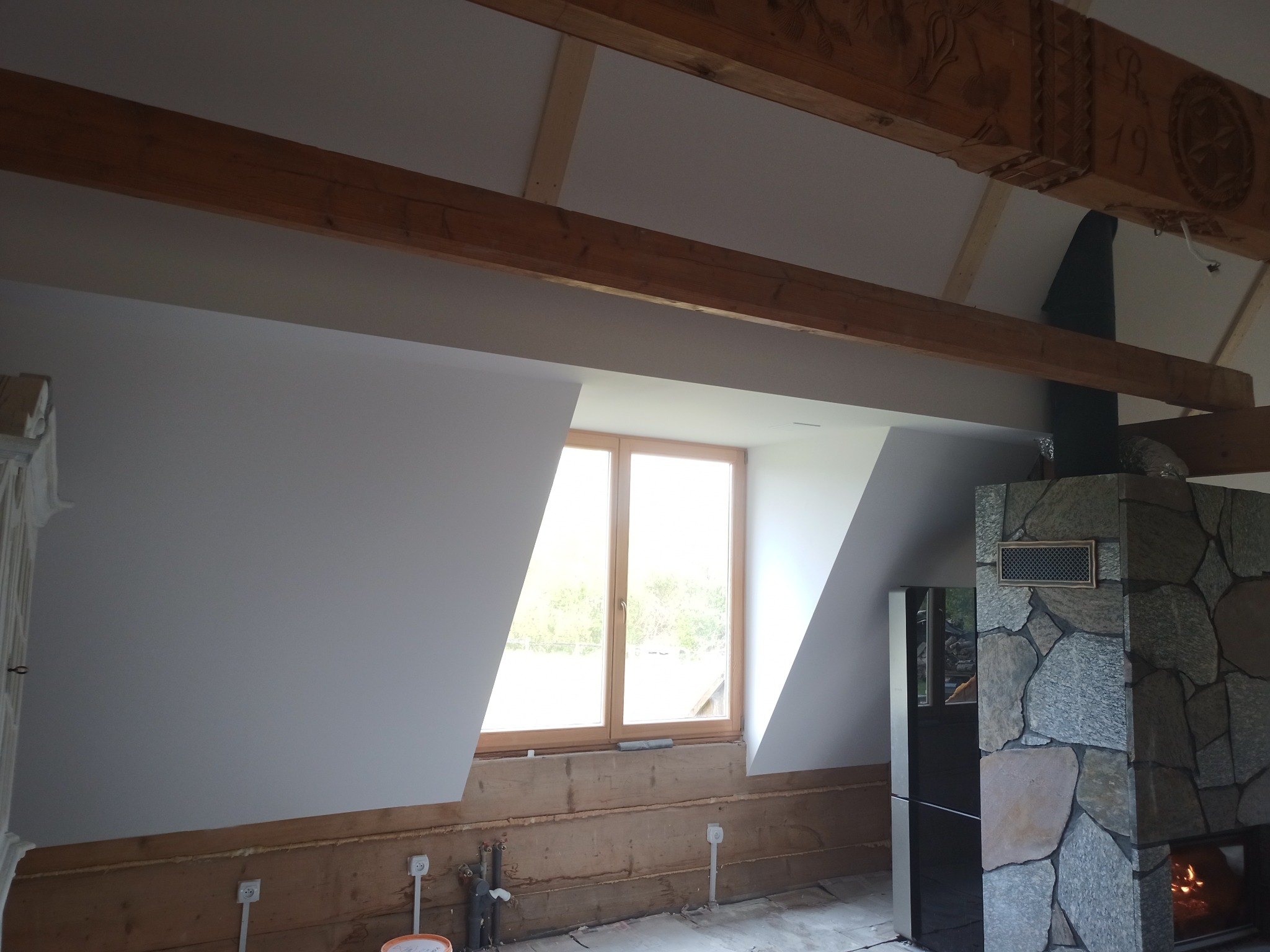 Poddasze w trakcie prac wykończeniowych: widoczne drewniane belki stropowe z dekoracyjnymi rzeźbieniami, gładkie, białe ściany skośne, okno w drewnianej ramie, fragment murowanego kominka z czarnym...