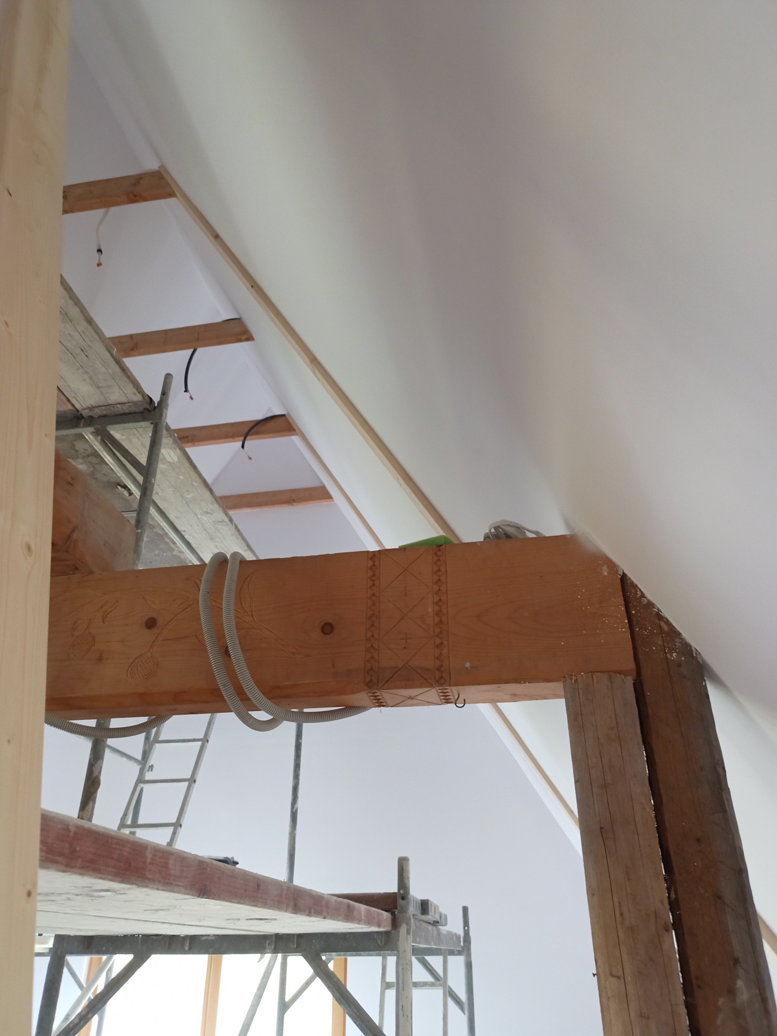 Poddasze w trakcie prac wykończeniowych: widoczne ocieplenie skośnego dachu, drewniana konstrukcja nośna z dekoracyjnymi rzeźbieniami oraz rusztowanie malarskie.