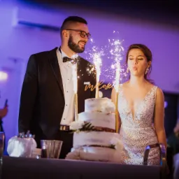 Uśmiechnięty pan młody w okularach patrzy na pannę młodą, która zdmuchuje iskry z tortu weselnego, w tle goście i fioletowe oświetlenie.