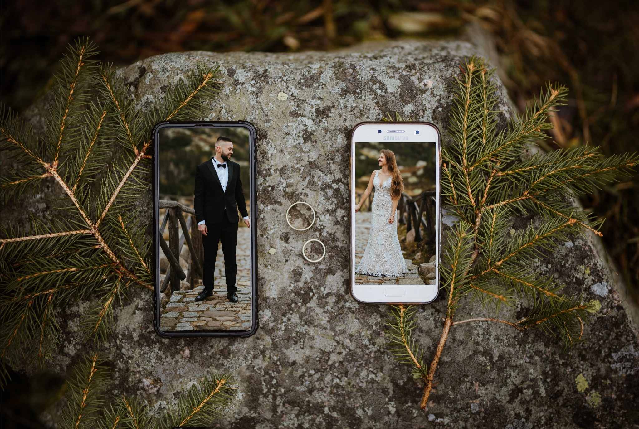 Dwa telefony komórkowe leżące na kamieniu, wyświetlające zdjęcia pary w strojach ślubnych, obok leżą obrączki, całość otoczona gałązkami świerkowymi.