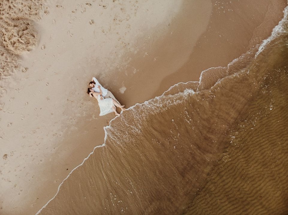 Para młoda leżąca na piaszczystej plaży, sfotografowana z lotu ptaka, z falami delikatnie obmywającymi brzeg.