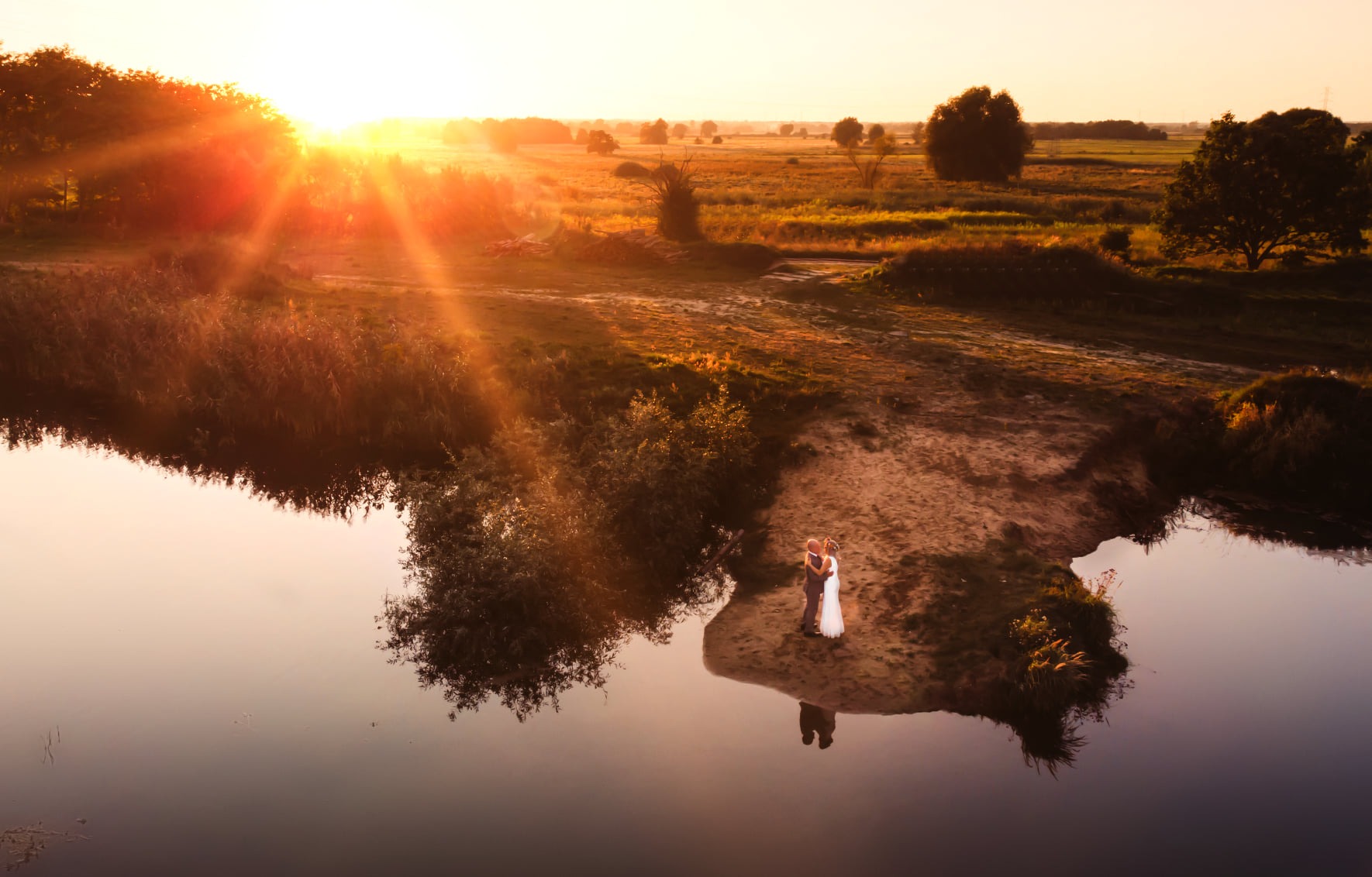 Para młoda w objęciach stoi na piaszczystym brzegu rzeki, odbicie w wodzie, zachód słońca w tle.