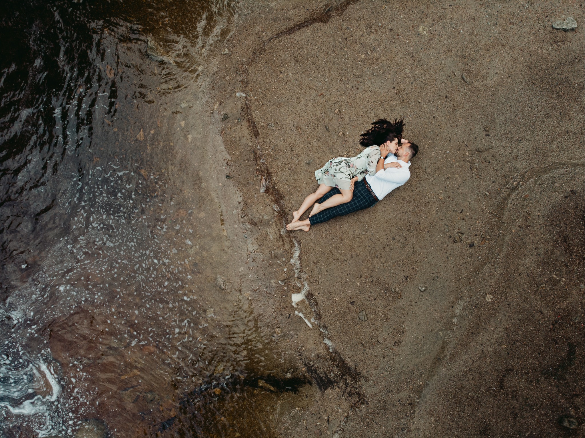Zakochana para leżąca na piasku obok brzegu wody, widok z góry, ona w kwiecistej sukience, on w białej koszuli i spodniach w kratę, delikatny pocałunek.