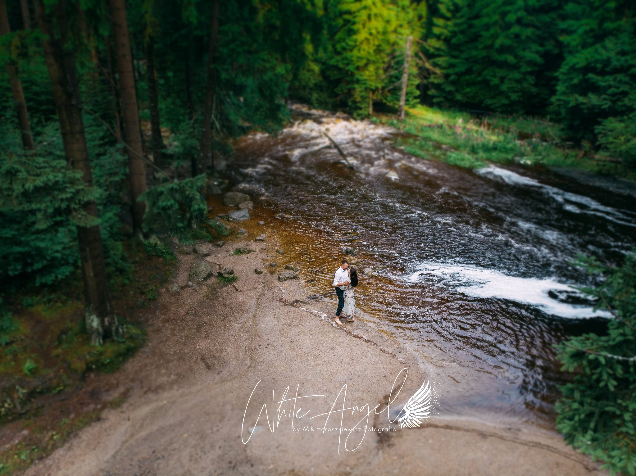 Para obejmująca się na piaszczystym brzegu rwącej rzeki w otoczeniu gęstego lasu, widok z góry, logo fotografa w dolnej części kadru.