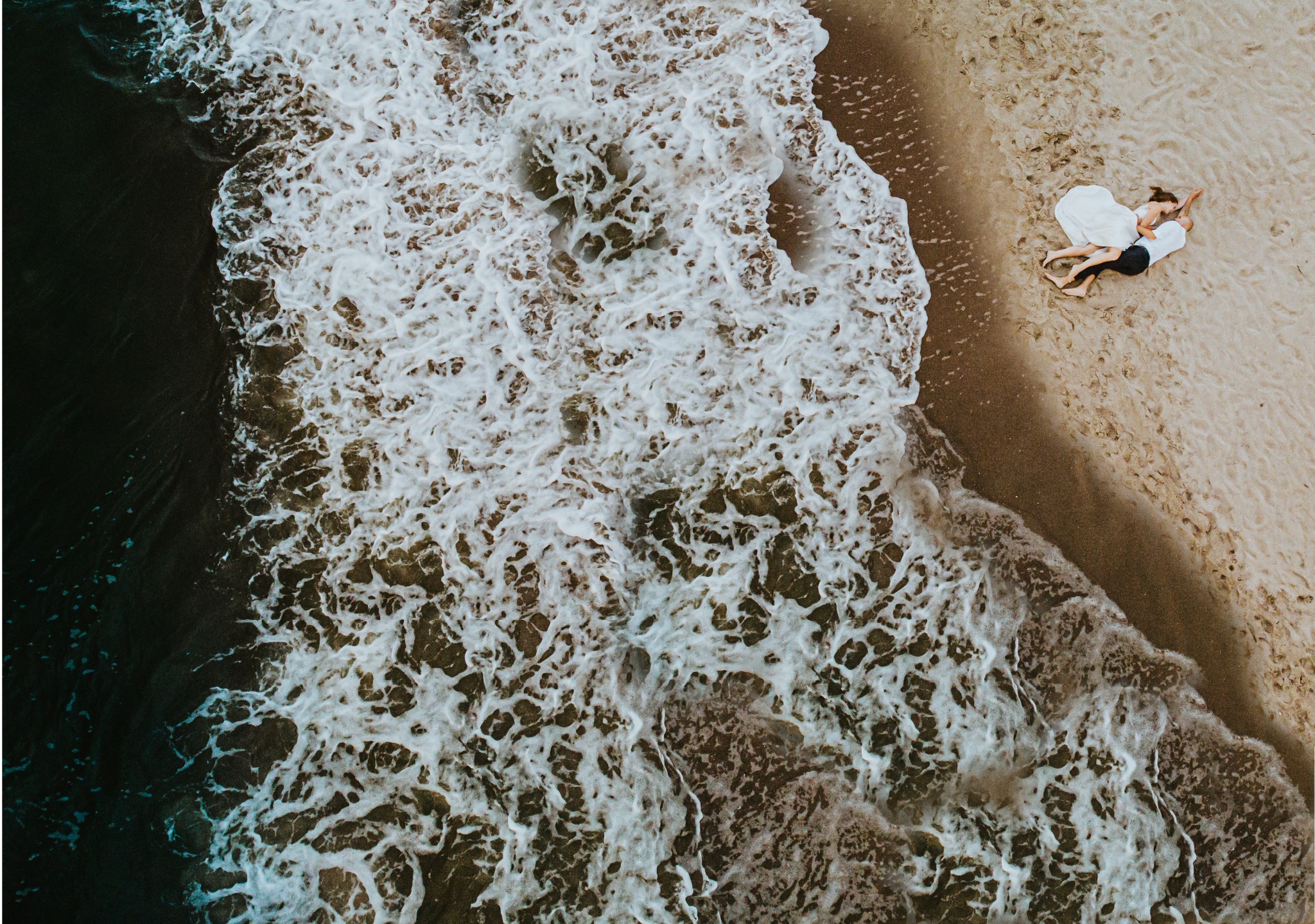 Widok z lotu ptaka na parę leżącą na piaszczystej plaży, tuż przy brzegu morza z burzliwymi falami.