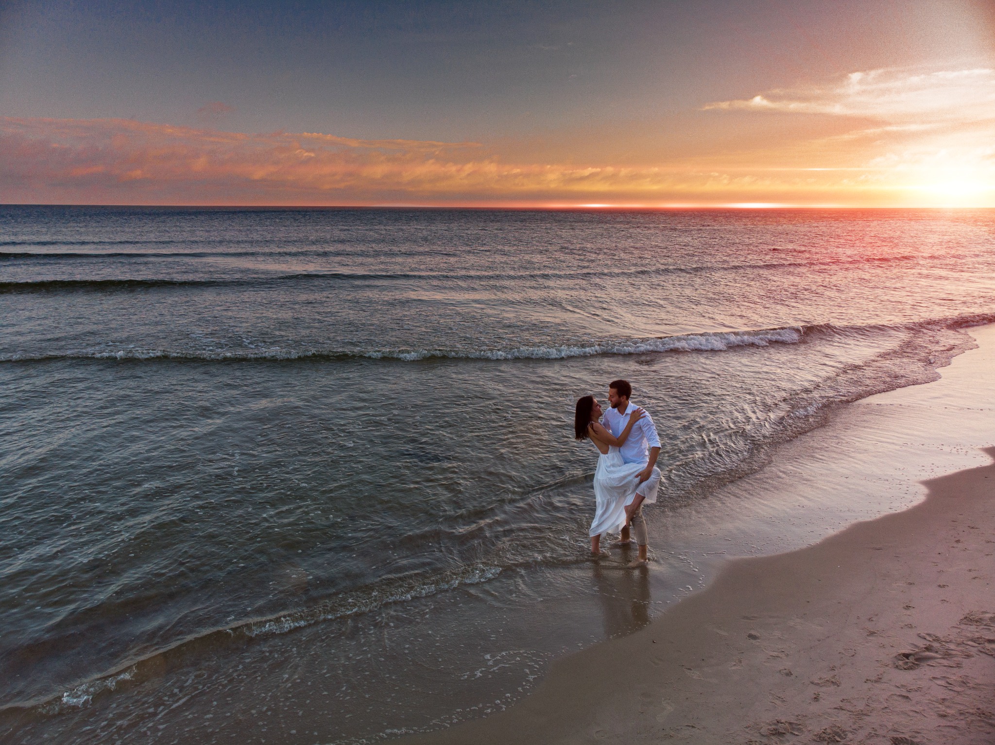 Para zakochanych na plaży o zachodzie słońca, mężczyzna trzyma kobietę w ramionach, oboje w białych ubraniach, morze i pomarańczowe niebo w tle.