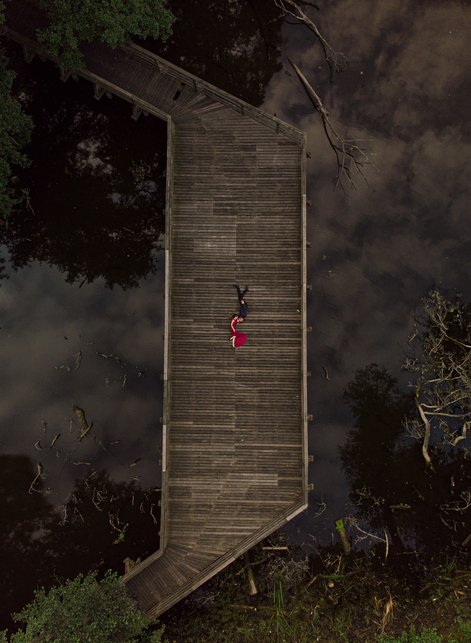 Para leży na drewnianym pomoście nad bagnem, widok z lotu ptaka, kobieta w czerwonej sukience.