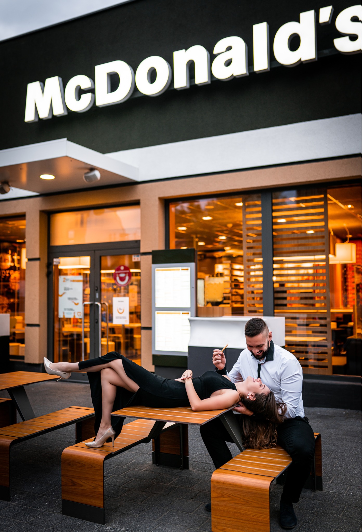 Uśmiechnięta para: kobieta w czarnej sukience leży na stole przed restauracją McDonald's, mężczyzna w białej koszuli siedzi obok, trzymając frytkę.
