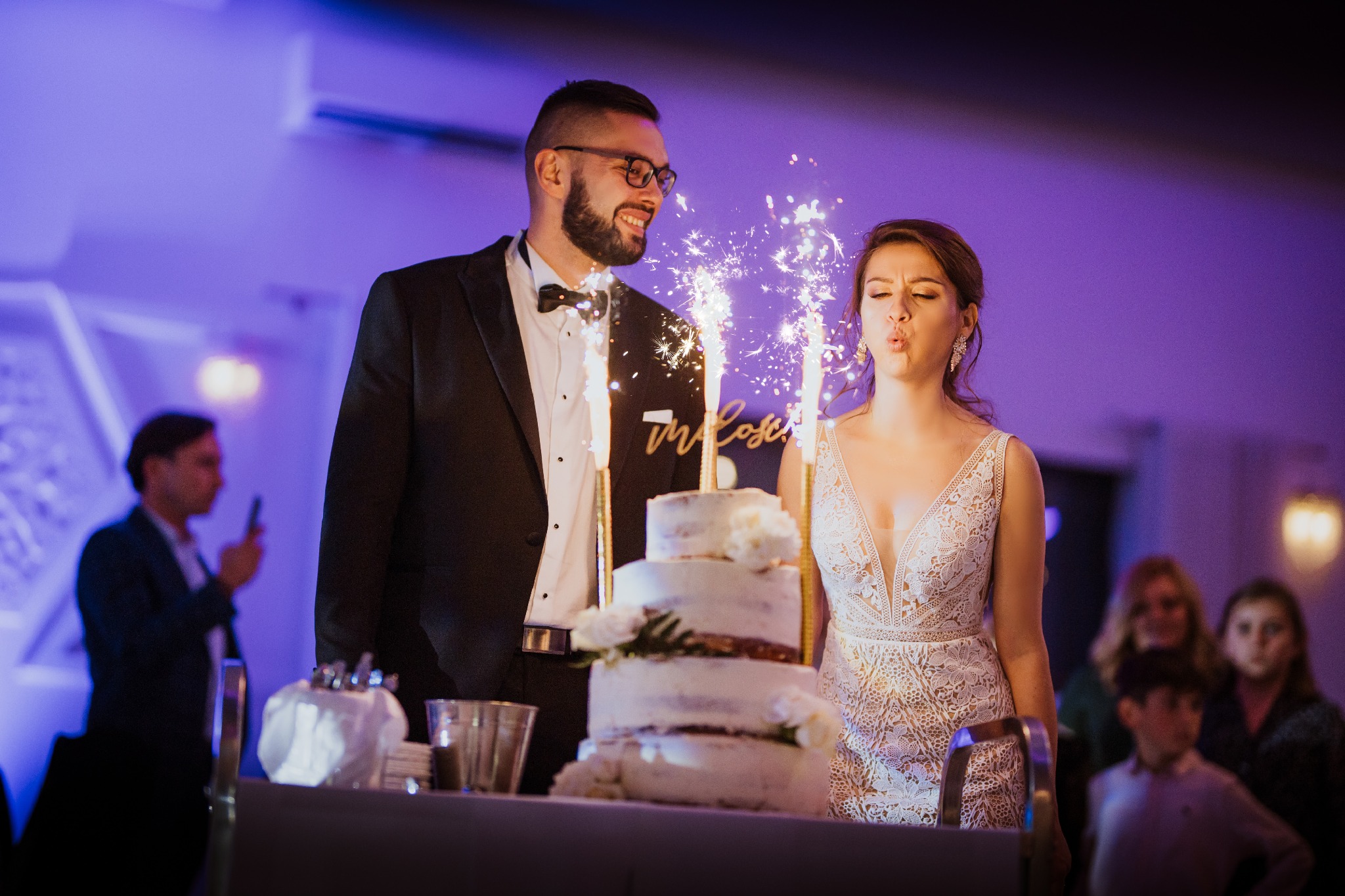 Uśmiechnięty pan młody w okularach patrzy na pannę młodą, która zdmuchuje iskry z tortu weselnego, w tle goście i fioletowe oświetlenie.