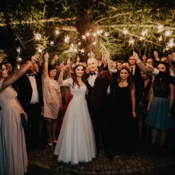 Para młoda otoczona gośćmi weselnymi trzymającymi zapalone zimne ognie pod rozświetlonym drzewem.