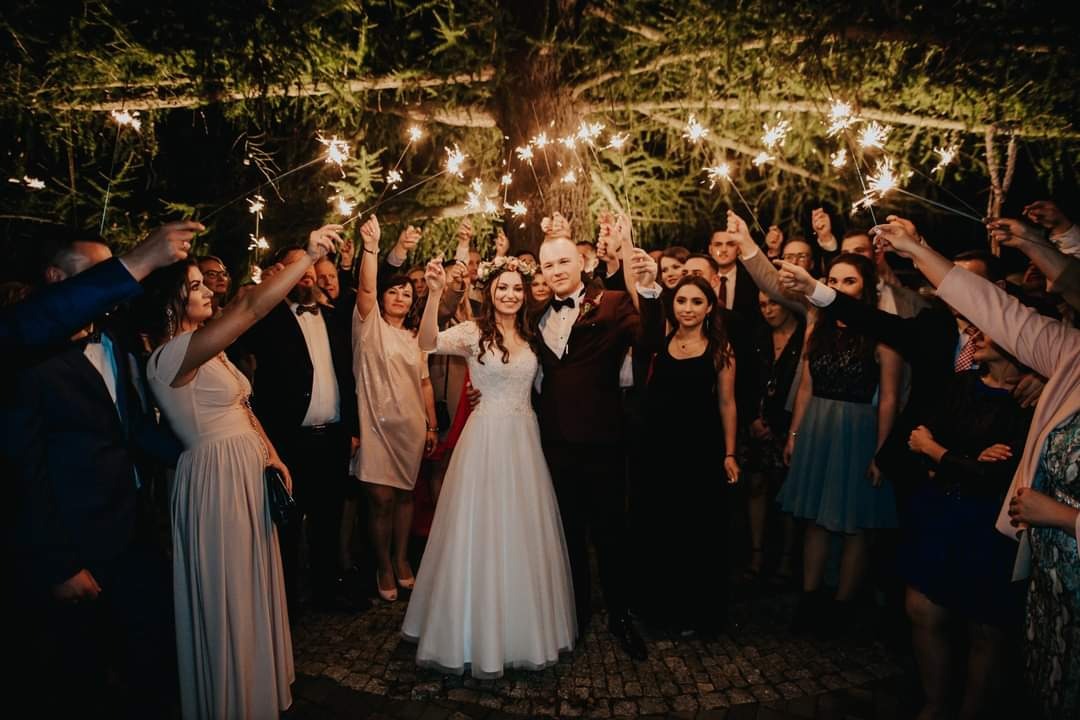 Para młoda otoczona gośćmi weselnymi trzymającymi zapalone zimne ognie pod rozświetlonym drzewem.