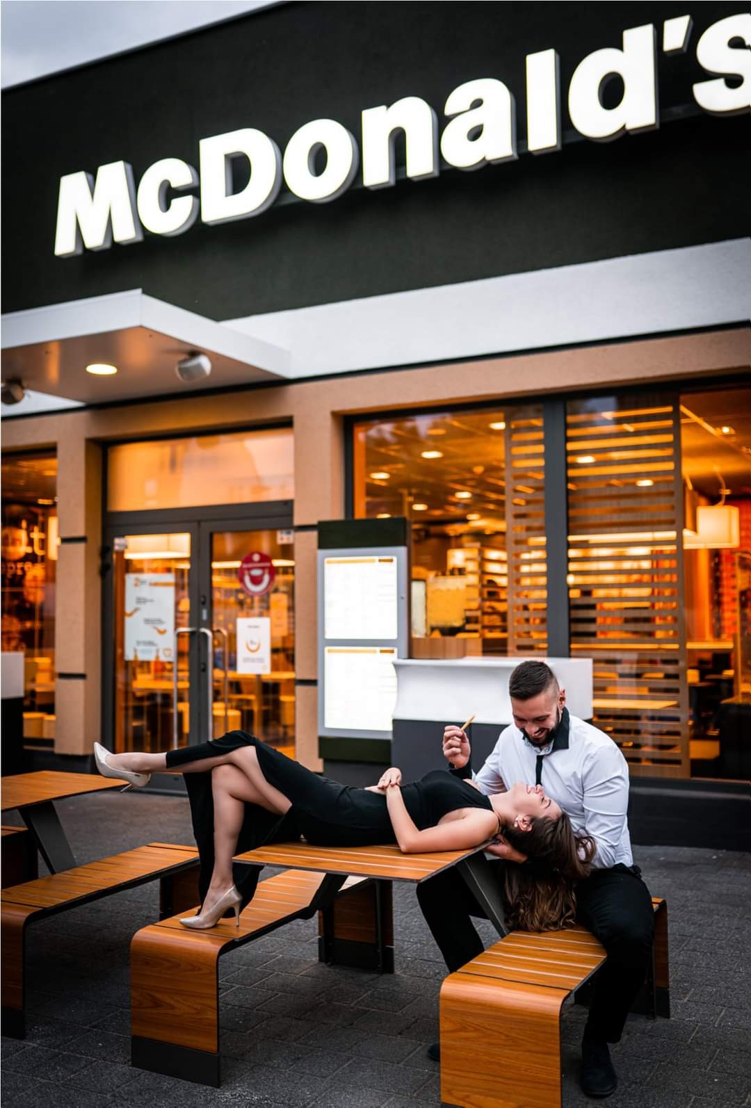 Para w eleganckich strojach pozuje przed restauracją McDonald's: kobieta leży na stole, mężczyzna siedzi obok trzymając frytkę.