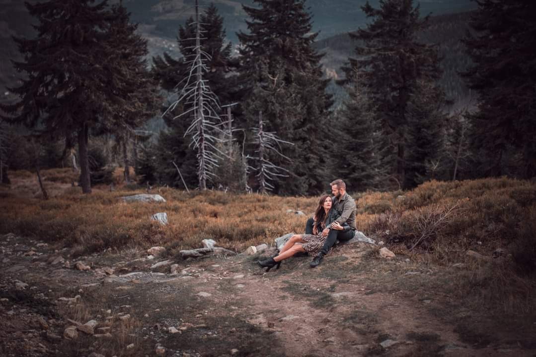 Para siedząca na kamieniu w górskim krajobrazie z lasem w tle, kobieta w sukience w panterkę i czarnych botkach, mężczyzna w zielonej koszuli, oboje patrzą w lewo.