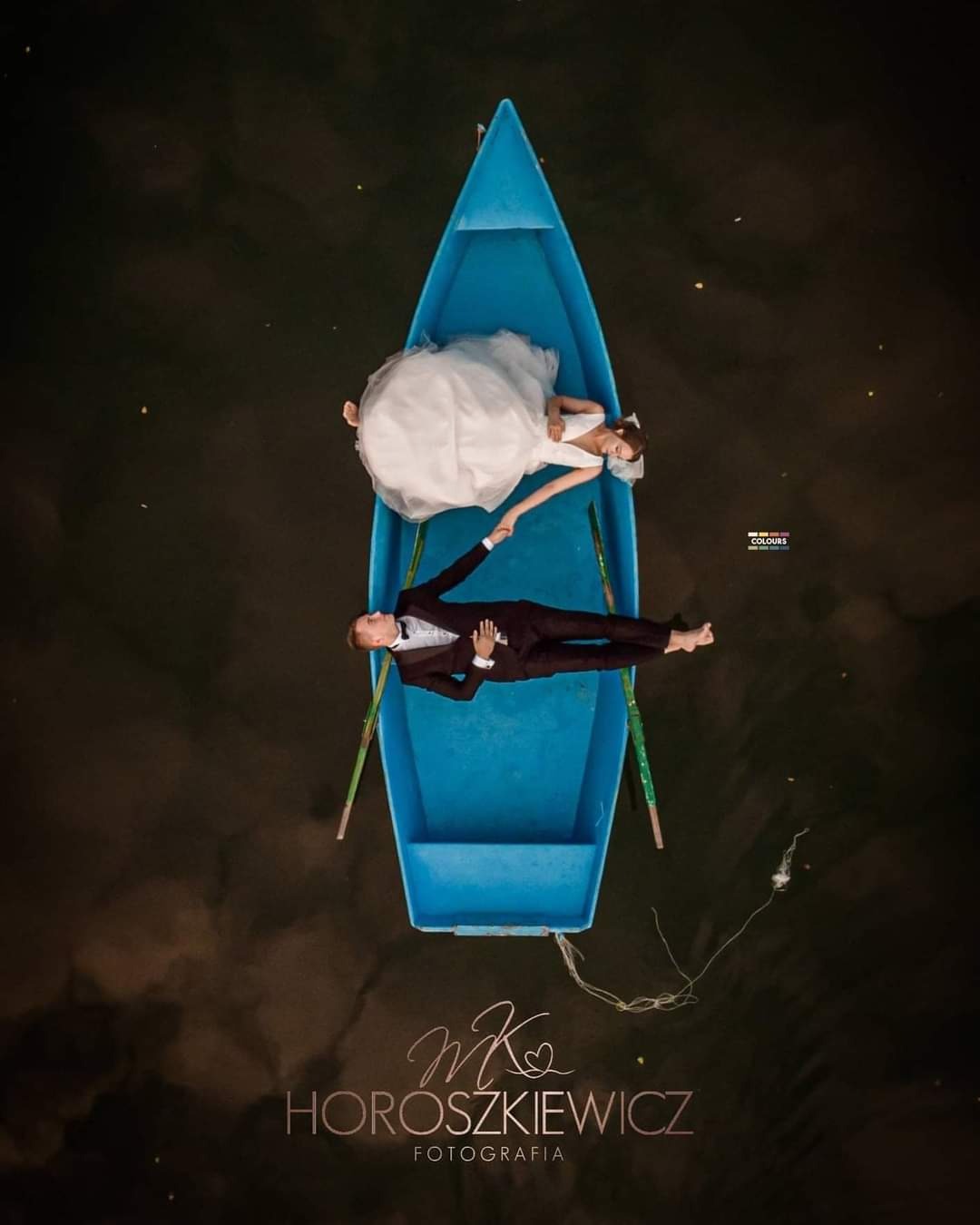 Zdjęcie z lotu ptaka przedstawia parę młodą leżącą w błękitnej łódce na ciemnej wodzie. Panna młoda w białej sukni i welonie, pan młody w ciemnym garniturze, trzymają się za ręce. W tle widoczne...