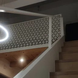 Drewniane schody z białą balustradą o geometrycznym wzorze, oświetlone nowoczesną lampą w kształcie okręgu, prowadzące na antresolę z drewnianym stropem.