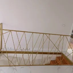 Złota balustrada schodowa z geometrycznym wzorem krzyżujących się pręt&oacute;w, widok z g&oacute;ry na marmurowe schody i fragment pokoju na poddaszu.