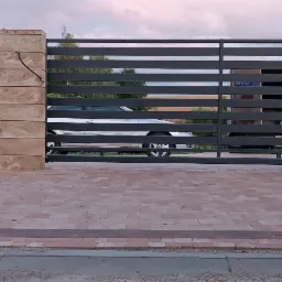 Nowoczesna brama wjazdowa przesuwna z poziomych profili metalowych, osadzona między słupkami z imitacji kamienia, widok z zewnątrz na podjazd z kostki brukowej.