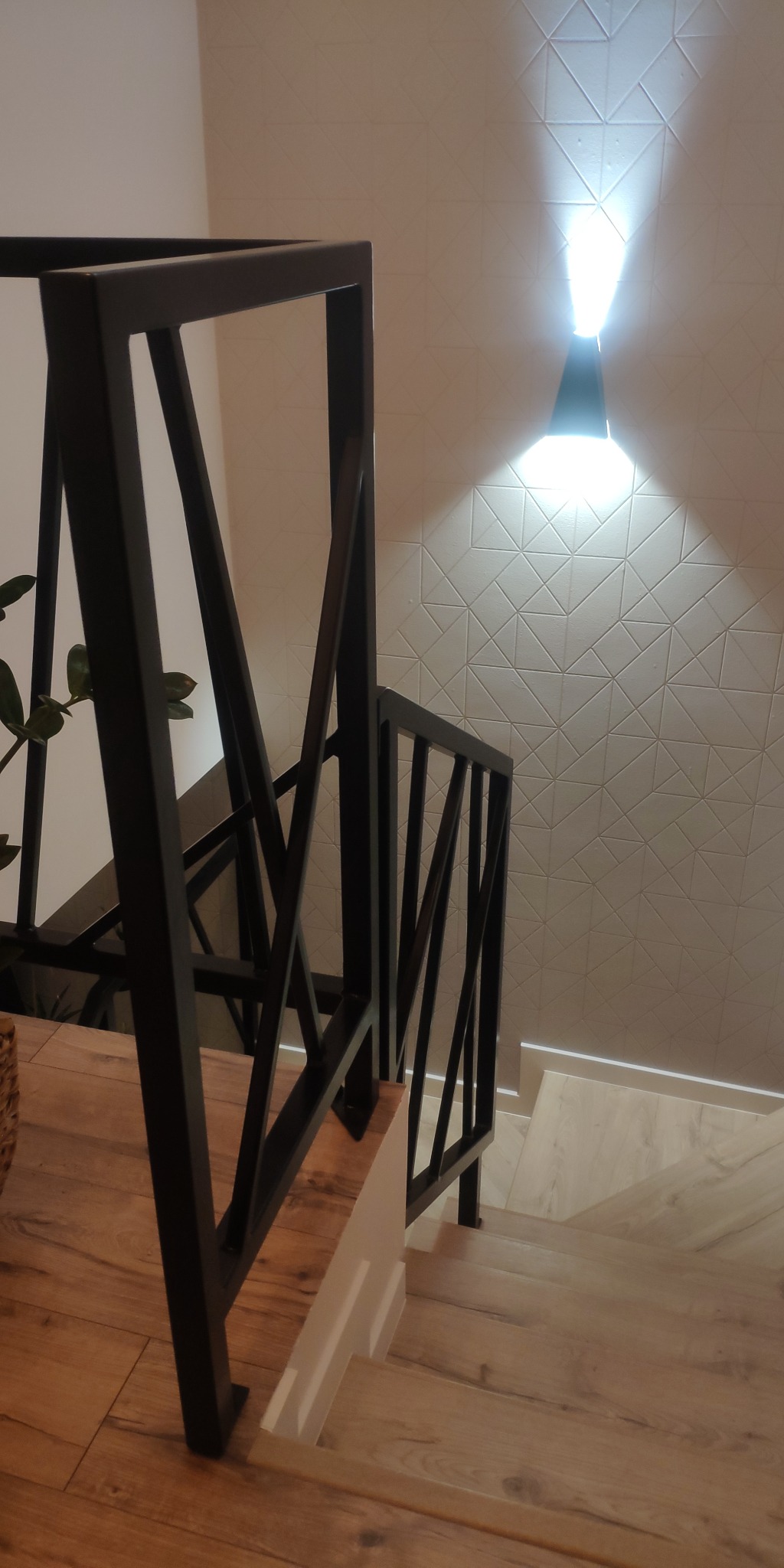 Nowoczesne schody z czarną, metalową balustradą o geometrycznym wzorze, oświetlone lampą ścienną z wzorem trójkątów na ścianie, widoczne z góry.