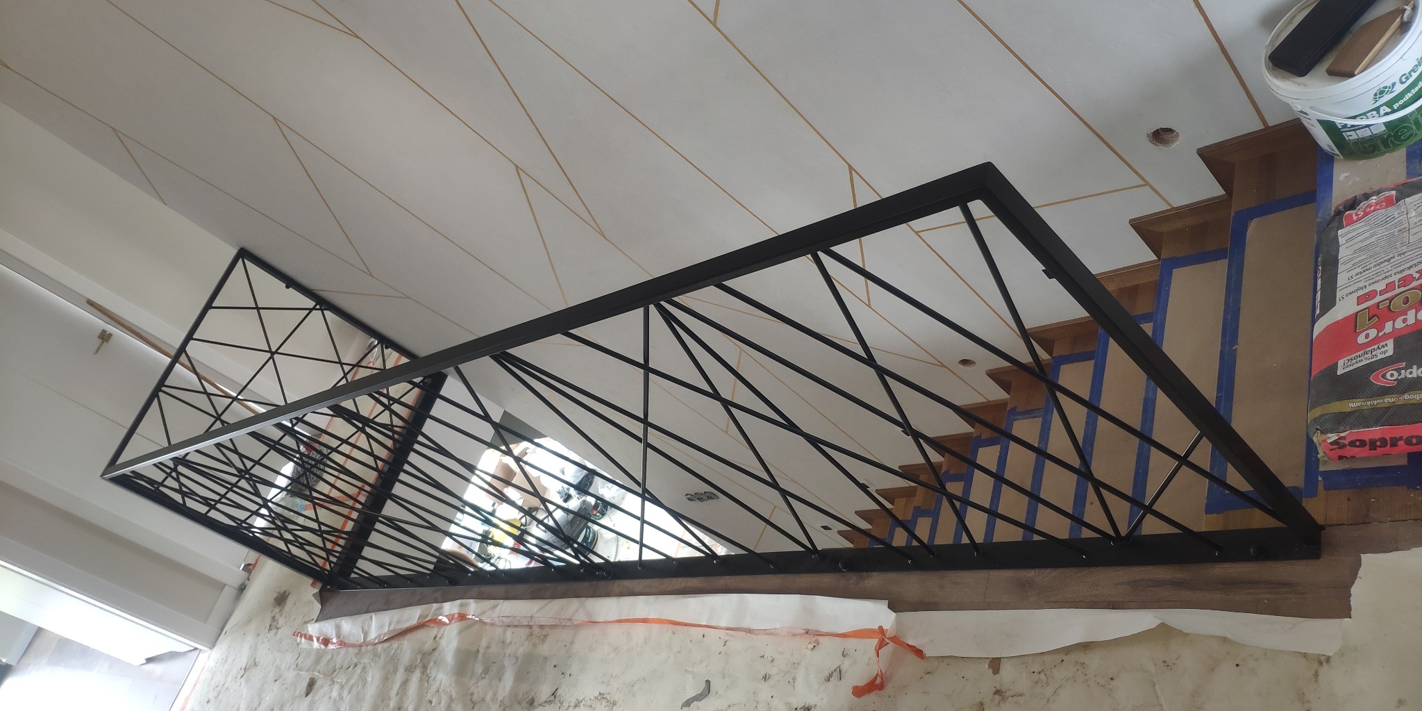 Nowoczesna balustrada metalowa o geometrycznym wzorze, zamontowana przy drewnianych schodach zabezpieczonych taśmą malarską w trakcie remontu w Białobrzegach. Widoczne materiały budowlane i narzędzia.