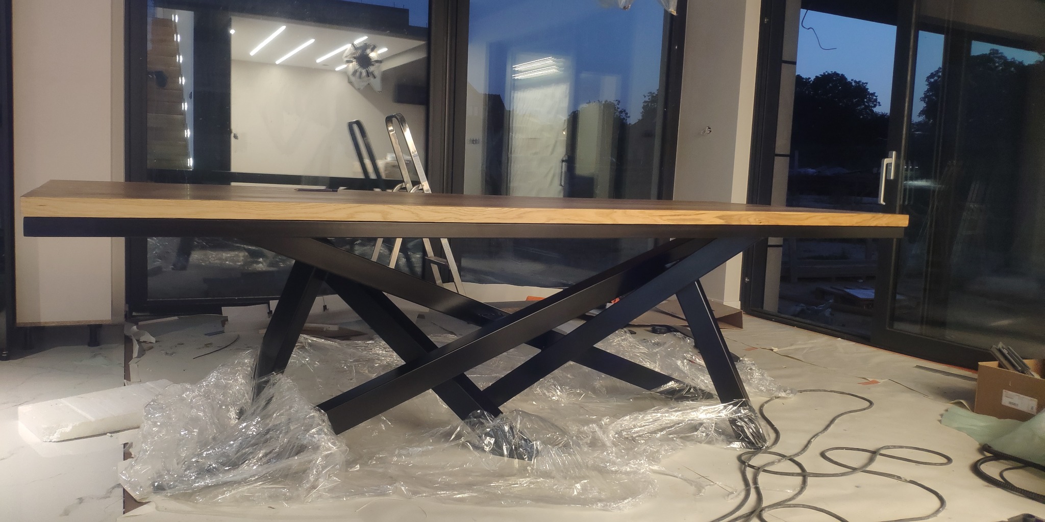Stół z drewnianym blatem i metalową podstawą w kształcie litery X, zabezpieczony folią ochronną podczas montażu, widoczne okna przesuwne w tle.