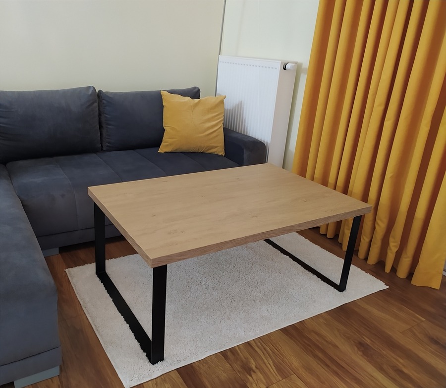 Stolik kawowy z blatem imitującym jasne drewno, na czarnych, metalowych nogach w salonie z szarą sofą narożną, żółtą poduszką i zasłonami w odcieniu musztardowym.