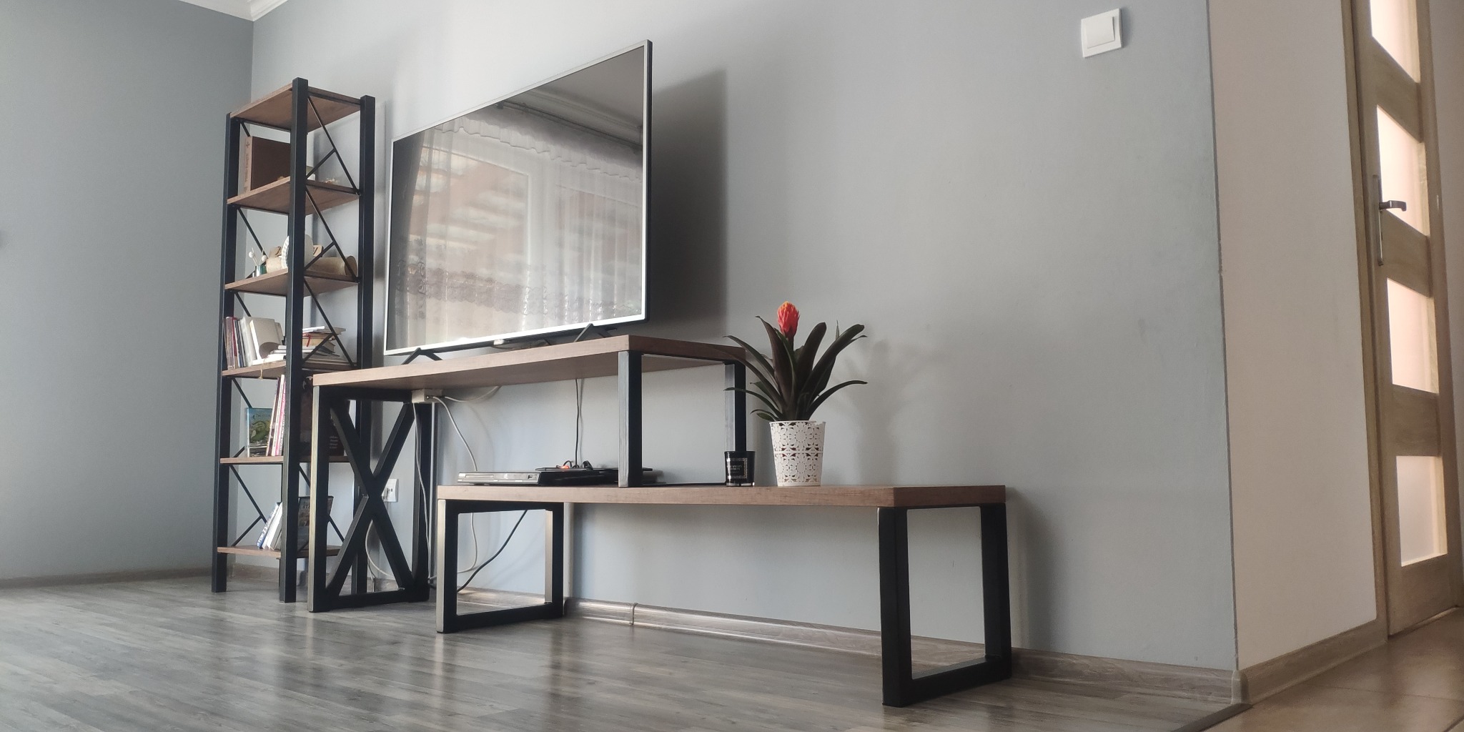 Regał i stolik RTV z metalowymi elementami w stylu loft, uzupełnione o dekoracje w postaci książek i rośliny doniczkowej, na tle szarej ściany i drewnianej podłogi.