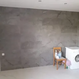 Wykończona łazienka z szarymi płytkami imitującymi marmur na ścianie, białą podłogą i pralką. Na krześle stoją środki czystości.