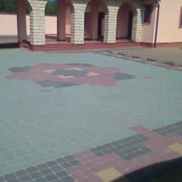 Brukowana nawierzchnia podjazdu z wzorem w kształcie rombu, widok na budynek z kolumnami i łukami, obrzeża wykończone kostką.