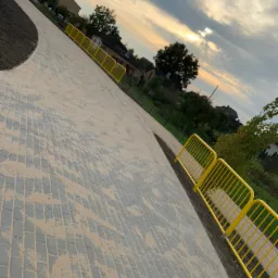 Nowo ułożona, zakrzywiona ścieżka z kostki brukowej w Płużnicy, zabezpieczona żółtymi barierkami, w tle zabudowa mieszkalna i zachodzące słońce.