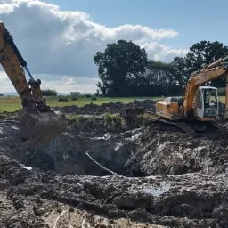 Dwa żółte koparki CAT i Liebherr pracują na polu, wykopując dół w błotnistej ziemi, w tle widoczne drzewa i niebo z chmurami.
