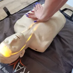 Ręce z niebieskim manicure wykonują masaż serca na fantomie na szarej macie treningowej z logo Grupy Poszukiwawczo-Ratowniczej LIFE.