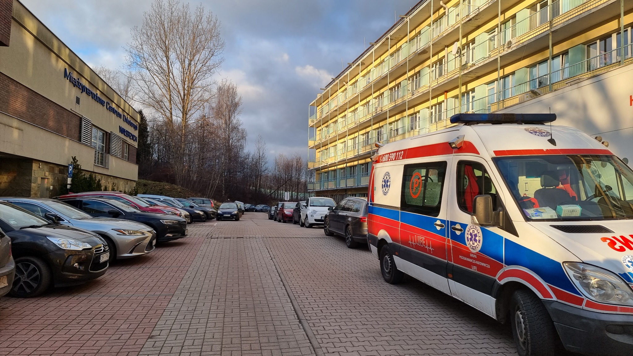 Ambulans zaparkowany przed budynkiem Międzynarodowego Centrum Okulistyki, z widocznymi samochodami osobowymi w tle.