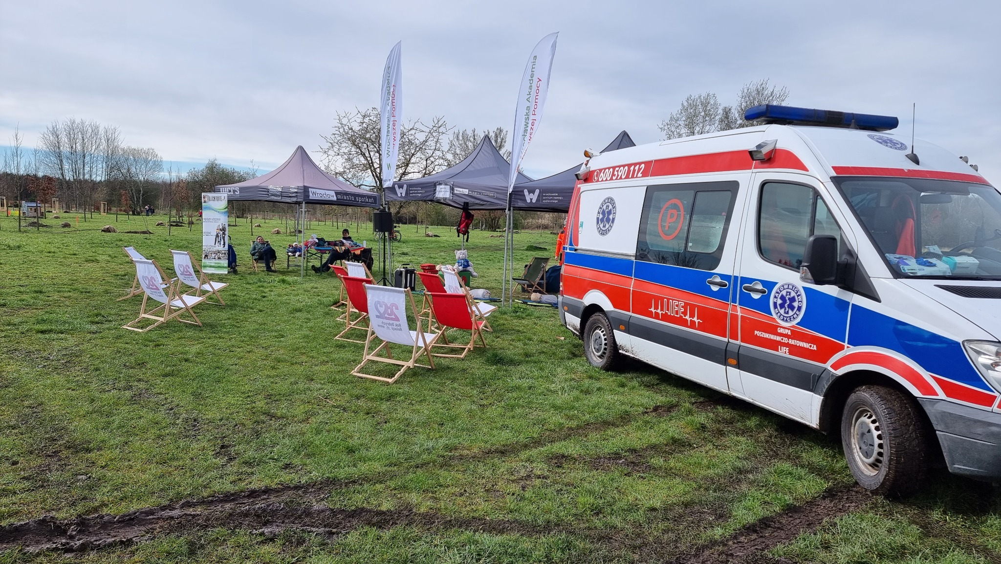 Ambulans zaparkowany na trawniku obok rozstawionych namiotów i leżaków z logo organizatora, prawdopodobnie podczas plenerowego pokazu lub szkolenia z zakresu ratownictwa.
