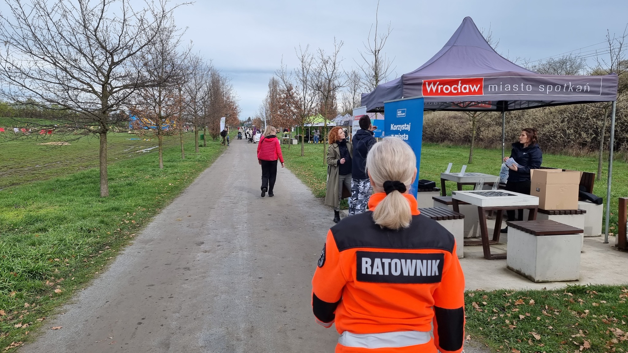 Ratownik medyczny w pomarańczowej kurtce z napisem 'RATOWNIK' na plecach, stojący na ścieżce w parku, w tle stoisko promocyjne miasta Wrocław.