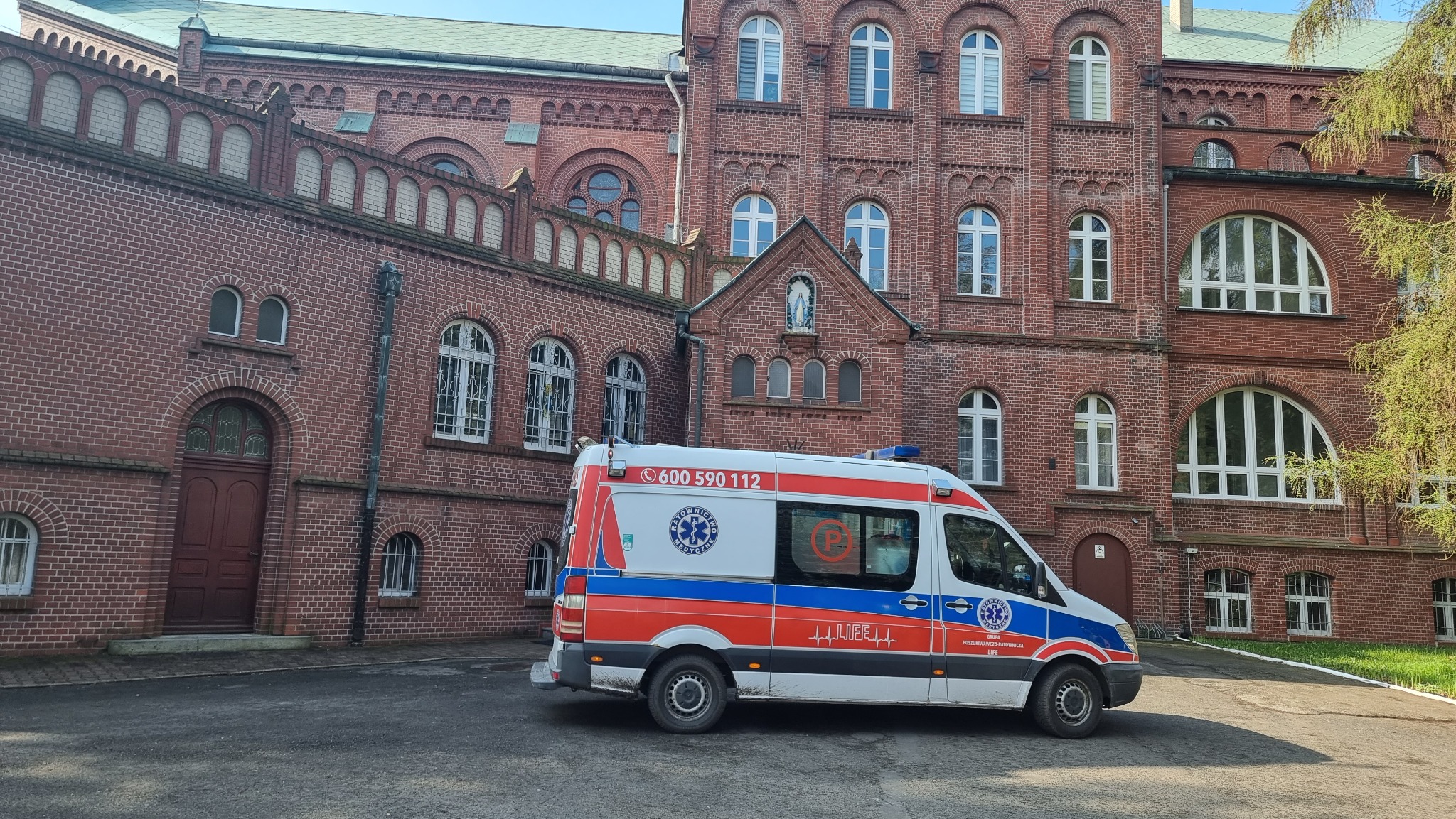 Ambulans zaparkowany przed budynkiem z czerwonej cegły z widoczną figurką Matki Boskiej