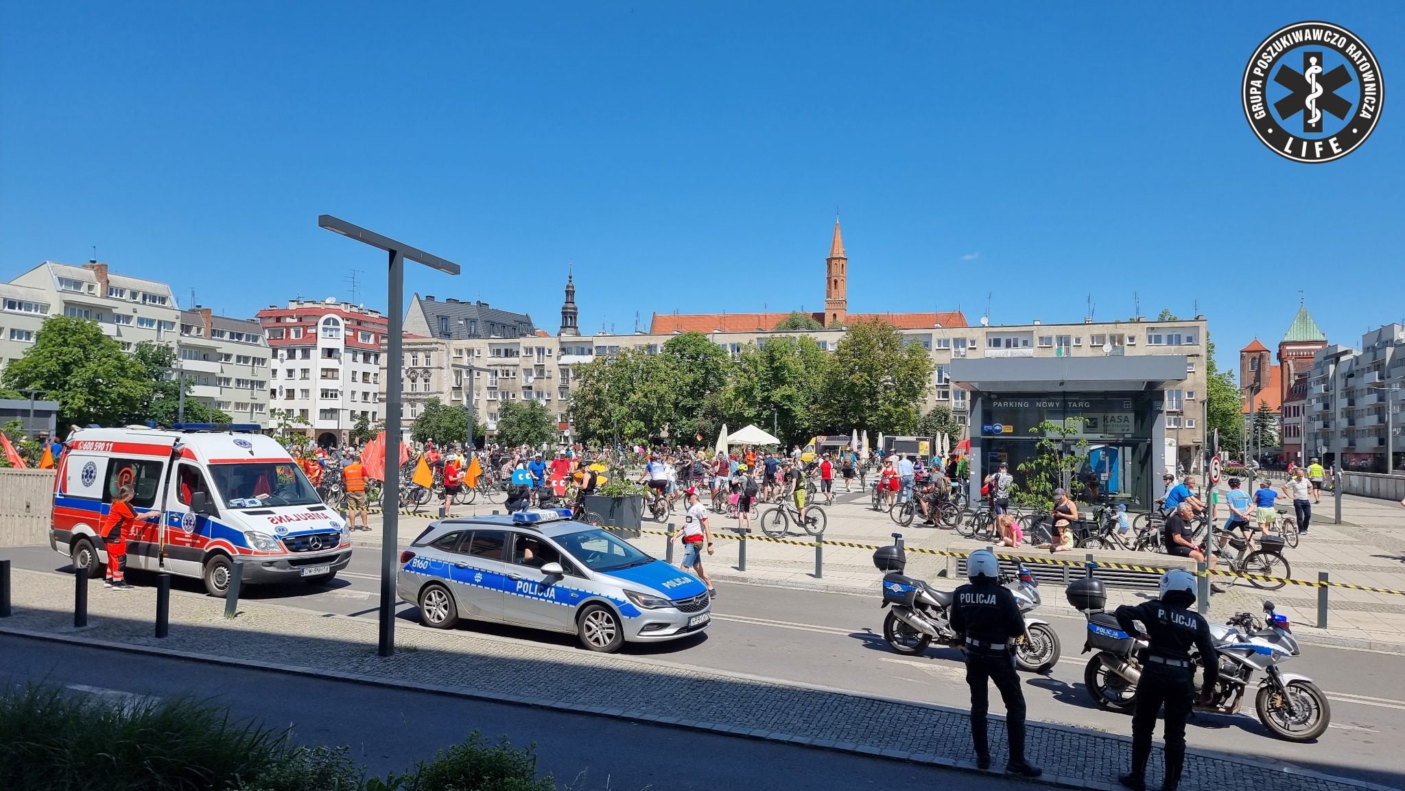 Ambulans, radiowóz policyjny i motocykle policyjne zabezpieczają rowerowy przejazd przez plac miejski z zabytkową zabudową w tle.