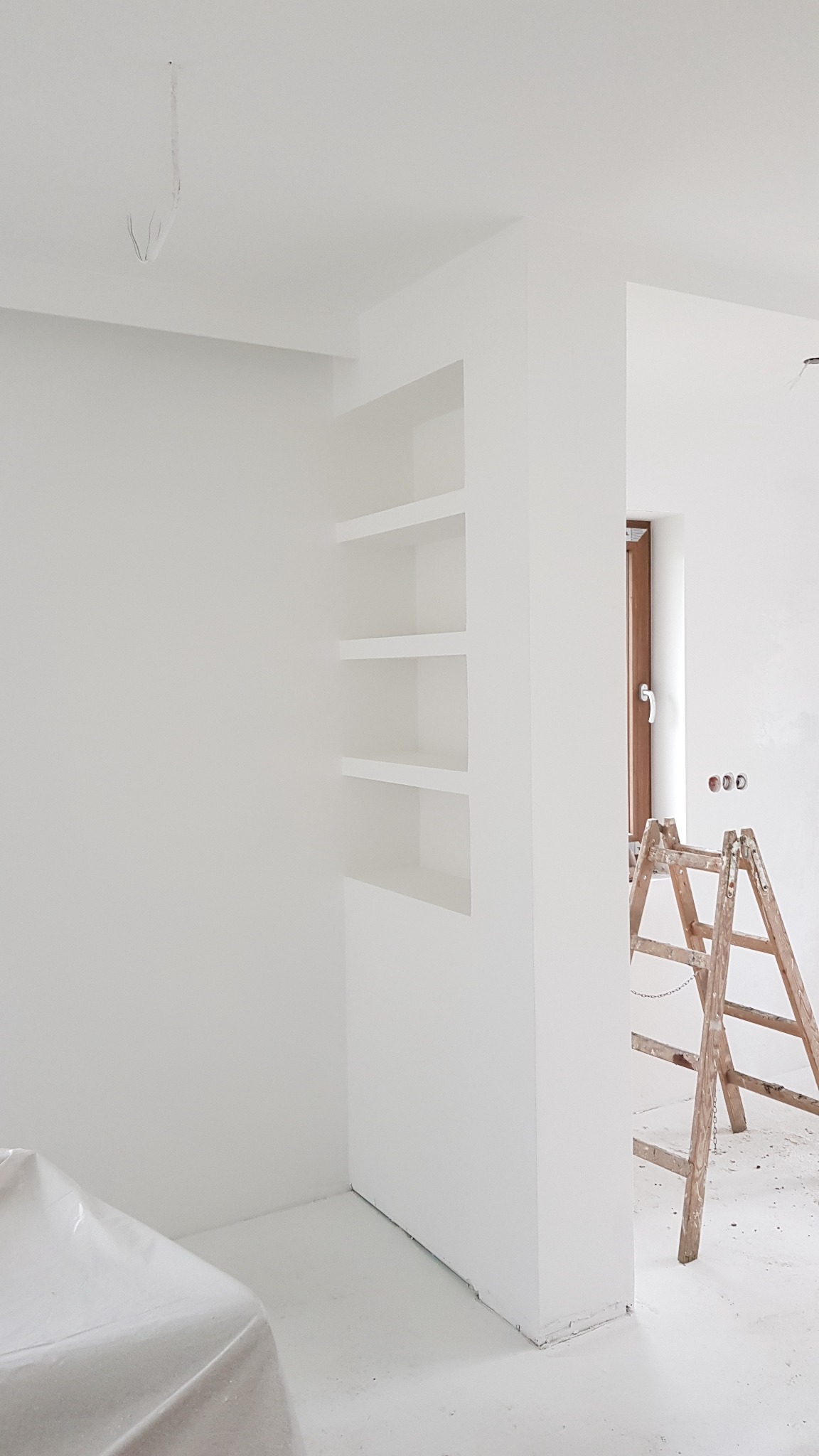 Białe wnętrze w trakcie remontu: pomalowane ściany, wnęka z półkami, drewniana drabina oparta o ścianę, zabezpieczone meble folią malarską, widoczne przewody elektryczne.