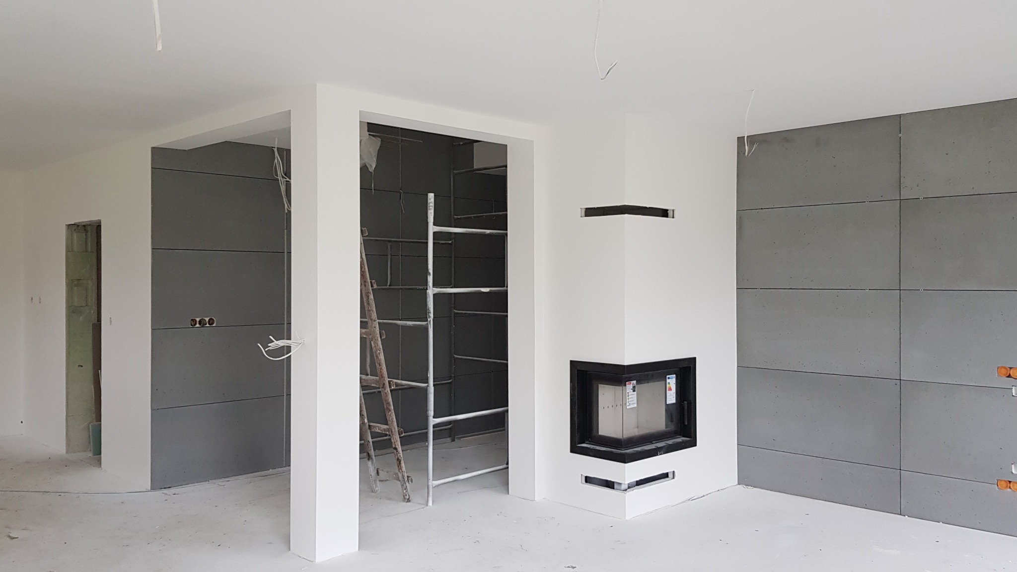 Wnętrze w trakcie prac wykończeniowych: ściany pokryte szarymi panelami imitującymi beton, biały kominek z czarnym obramowaniem w rogu, drewniana drabina oparta o ścianę, widoczne kable i puszki...