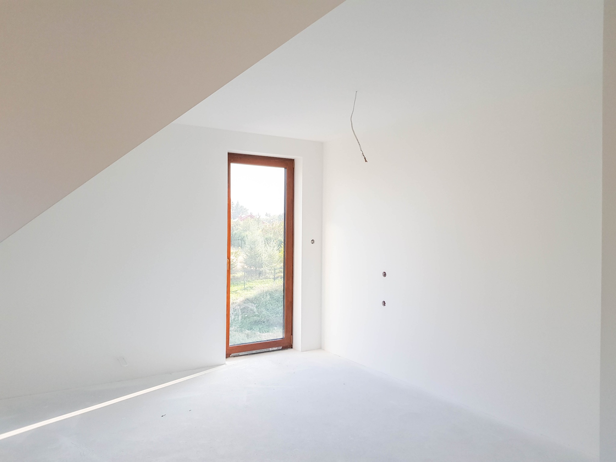 Pokój na poddaszu po świeżym pomalowaniu na biało, z drewnianym oknem i widocznymi śladami po instalacji elektrycznej.