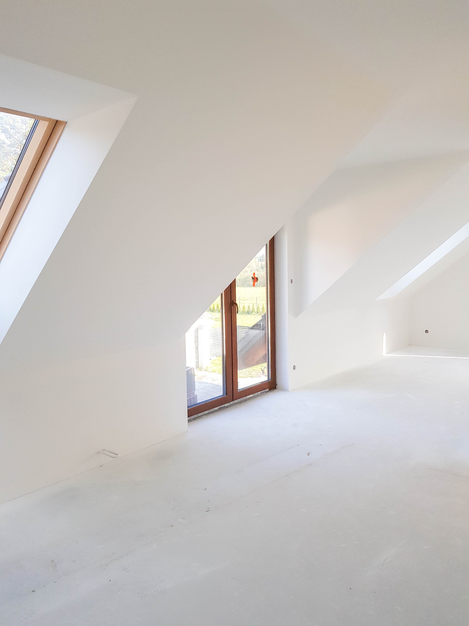 Poddasze po szpachlowaniu i gruntowaniu, widoczne okna dachowe i okno balkonowe, ściany i podłoga w jasnym odcieniu, przygotowane do dalszych prac wykończeniowych.